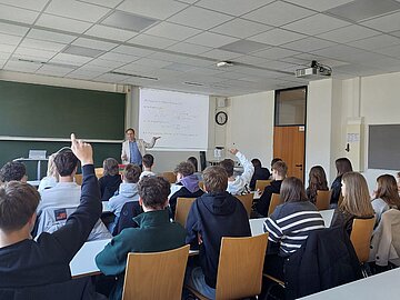 Gruppe von Schüler*innen von hinten zu sehen in einem Hörsaal sitzen, zwei davon melden sich. Vor ihnen ein Professor, der an einer Präsentation mit Mathematikaufgaben erklärt. 