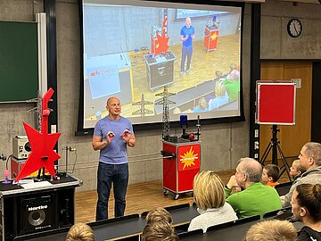 Mensch in einem Hörsaal, neben ihm rot-schwarze Experimenten-Tische, z. B. mit einer roten Bass-Gitarre. Im Hintergrund eine Leinwand mit dem selben Menschen in Vergrößerung. 