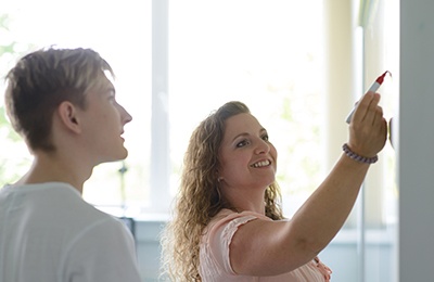 Zwei Studierende betrachten ein Whiteboard, sie schreibt etwas darauf, er schaut zu