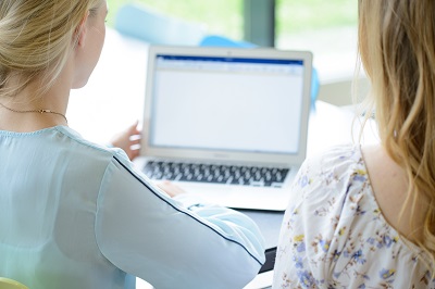 Blick von hinten auf zwei Studentinnen, die an einem Tisch sitzen und gemeinsam auf einen Laptop schauen, der in der Mitte des Bildes zu sehen ist. 