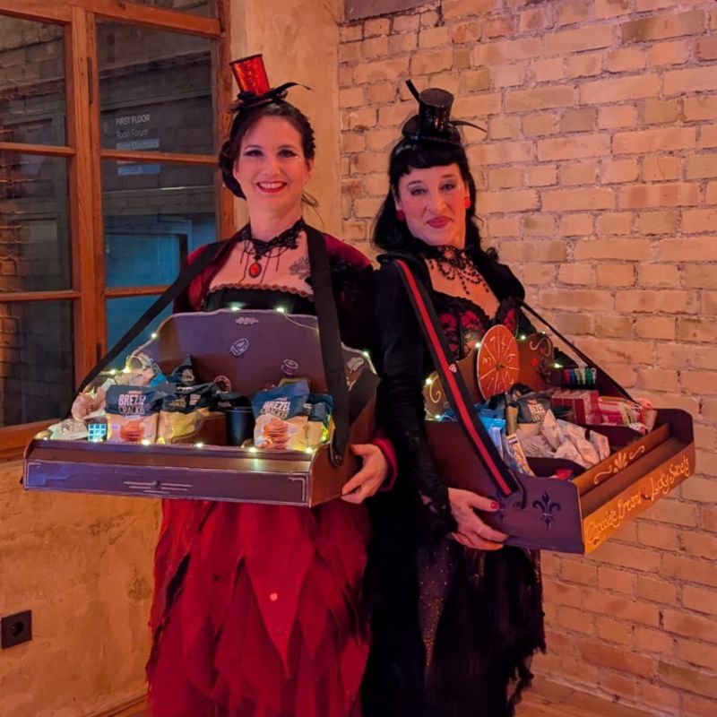 Zwei Frauen Rücken an Rücken, beide mit nostalgischen Kostümen in rot-schwarz und Bauchladen mit Snacks