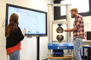 Im Labor Werkstoffprüfung der DHBW Mannheim: Ein Student (von vorne/seitlich zu sehen) bedient ein Universalprüfgerät, eine Studentin (von hinten zu sehen) steht vor einem großen Monitor mit Ergebnissen aus der Material-Prüfung