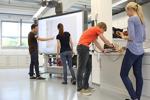 Grundlagen der Elektronik / Elektrotechnik festigen: Vier Studierende üben im Grundlagenlabor des Studiengangs Mechatronik an der DHBW Mannheim. Eine Gruppe (ein Student und eine Studentin) stehen an einem großen Monitor, er zeigt darauf. Die zweite Gruppe (ebenfalls ein Student und eine Studentin) bedient einen Multimeter für Spannungs- und Strommessungen