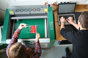 Montagewerkstatt des Studiengangs Mechatronik an der DHBW Mannheim, Ansicht von oben: Eine Studentin steht vor der Laserschneidmaschine und misst mit einem Messschieber eine gravierte, handgroße Glasplatte mit DHBW-Mannheim-Gravur, der daneben stehende Student tippt auf einen Laptop die zu gravierenden Informationen.