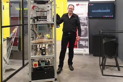 Mann an einem Messestand, links neben ihm steht der mannshohe Elektrochemische Verdichter in einem Aluminium-Profilrahmen. Er besteht aus mehreren Ebenen: Oben befindet sich ein zylindrischer Druckbehälter, in der Mitte ein Bedienpanel und darunter verschiedene Rohrleitungen, Pumpen sowie elektronische Steuerungskomponenten.