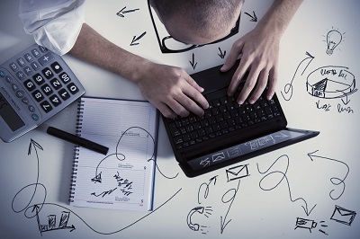 Mann von oben am Schreibtisch mit Händen auf Laptop-Tastatur. Auf dem Schreibtisch ein Taschenrechner und Notizblock, dazu Grafiken auf dem Schreibtisch.