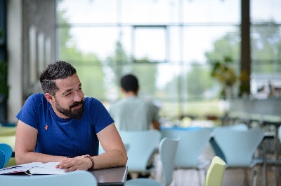 Mann mit dunklen Haaren und Bart in blauem T-Shirt in der DHBW-Mannheim-Mensa über einem Buch sitzend, schaut lächelnd nach rechts aus dem Bild heraus.