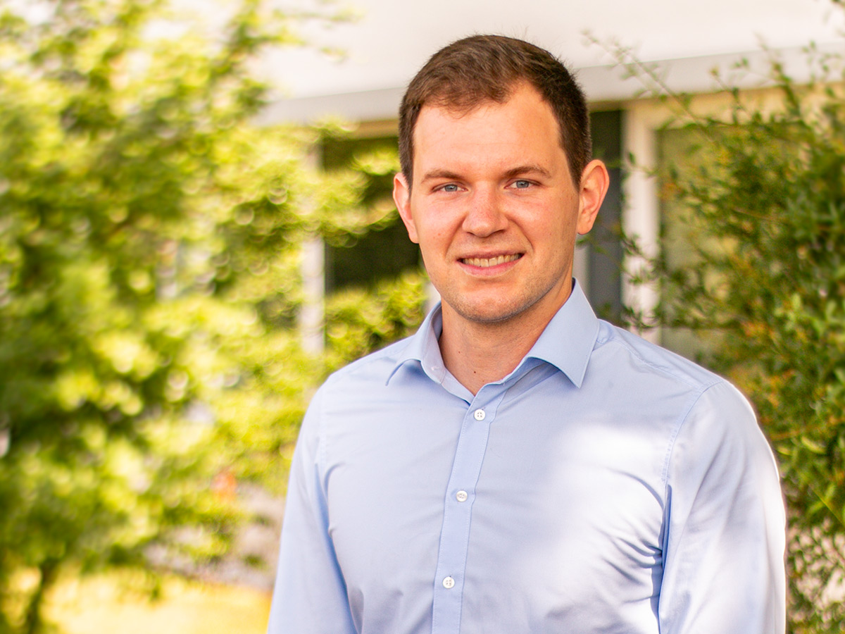Brustbild: Prof. Nathan Sudermann-Merx steht in hellblauem Hemd im Sonnenschein vor einem DHBW Gebäude im Grünen