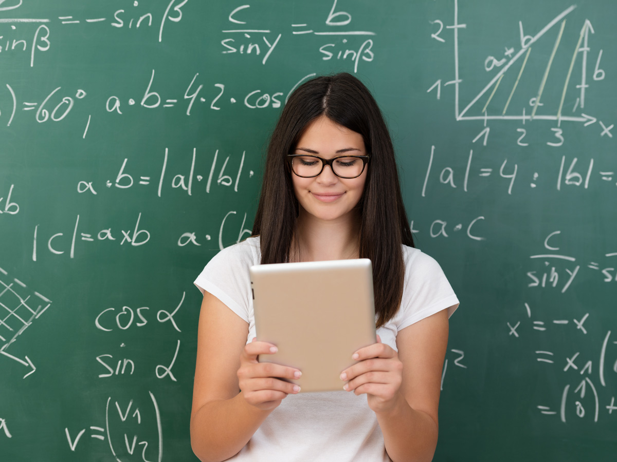 Junge Frau schaut auf ihr Tablet, im Hintergrund grüne Tafel mit mathematischen Gleichungen.