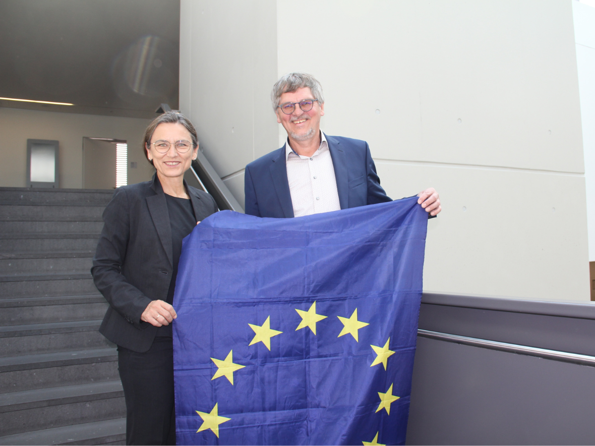 Die Präsidentin der DHBW neben einem Herrn im DHBW-Gebäude, gemeinsam halten sie die Flagge der EU.