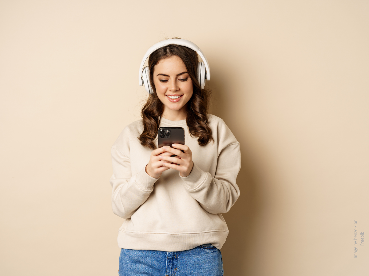 Junge Frau mit Kopfhörern schaut auf ihr Smartphone und lacht. Hintergrund beige, ihre Kleidung besteht aus einer Jeans und einem beigen Pullover. 