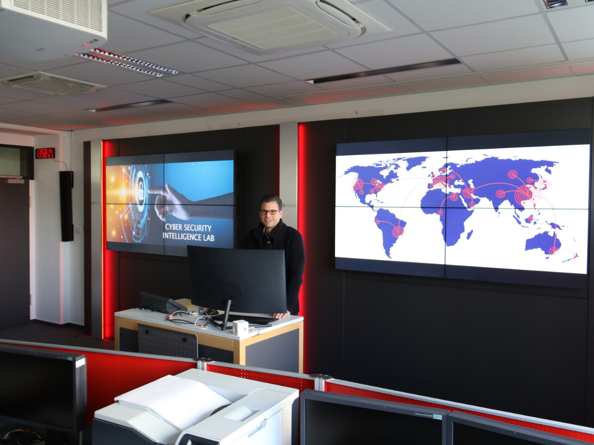 Prof. Bayreuther am Pult des Cyber Security Intelligence Labs vor zwei Monitor-Wänden.