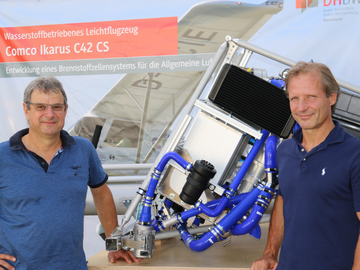 Porträt von Axel Schölch (li.) und Prof. Dr. Sven Schmitz, dazwischen der Versuchsaufbau EBSAL und im Hintergrund das Bild des Ultraleichtflugzeugs COMCO IKARUS C42 epower, auf das das EBSAL-Brennstoffzellensystem angepasst ist.  