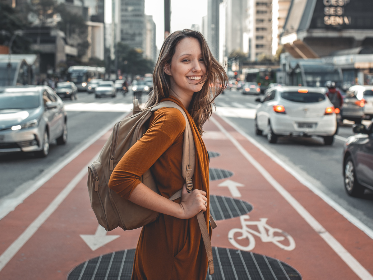 Junge Frau mit Rucksack steht auf einem Fahrradweg, links und rechts daneben Fahrstreifen mit Autos. Sie lächelt zuversichtlich in die Kamera.