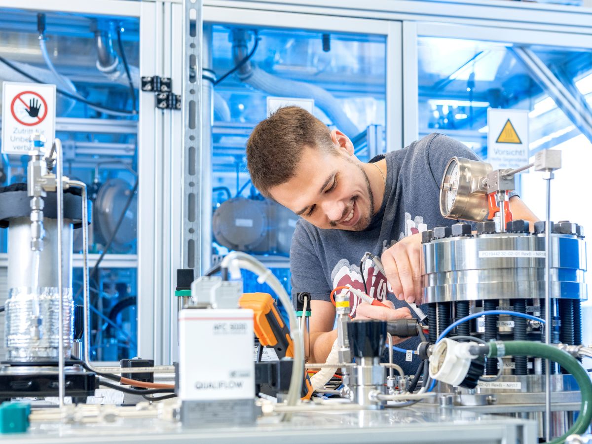 Porträt eines lachenden Mannes Mitte zwanzig im T-Shirt, der in einem Labor an einem 30 cm hohen, zylinderförmigen Elektrochemischen Verdichter lötet. 