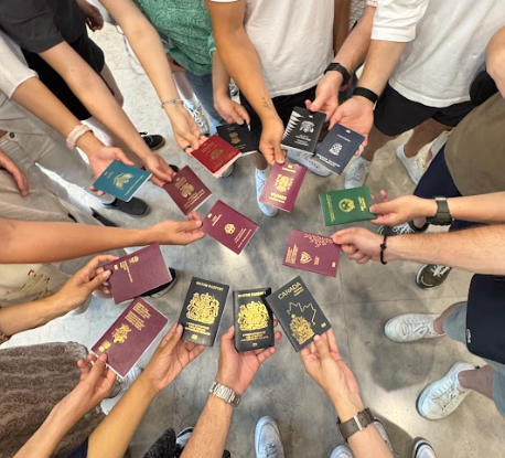 Bild von oben auf 15 Hände von im Kreis stehenden Studierenden, die ihre Pässe in die Mitte halten. 